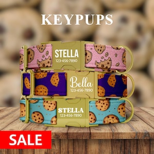 May include: Three personalized dog collars with cookie patterns in pink, purple, and teal. Each collar features a name and phone number on a gold-colored buckle. The word "KEYPUPS" is at the top, and "SALE" is at the bottom.