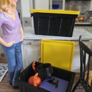 May include: A black storage bin with a yellow lid, containing miniature items. Inside are orange pumpkins, a black cauldron, and a purple book with a black silhouette. A doll stands nearby in a purple top and jeans.