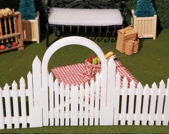 May include: White picket fence with a gate featuring an arched top. The fence is made of plastic and is approximately 12 inches tall. The gate is open and there is a red and white checkered picnic blanket behind it.
