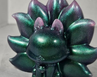 May include: A green and purple resin figurine of a stylized fox with nine tails. The fox has a cute, rounded design with small ears and a bell around its neck. The tails are large and fan out behind the fox.