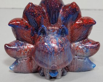 May include: A handcrafted figurine of a nine-tailed fox, featuring a vibrant blend of red, blue, and silver glitter. The sculpture has a smooth, glossy finish and a stylized, cartoonish design. The fox's tails are fanned out, creating a dynamic shape.