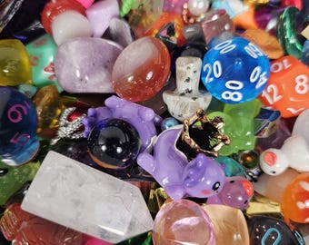 May include: A colorful assortment of gaming dice, gemstones, and small figurines. The collection includes various shapes and colors, such as blue, purple, orange, and green. There are also clear quartz crystals and a small gold-colored crown.