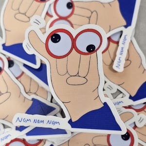 May include: A sticker featuring a cartoon hand with googly eyes wearing red-rimmed glasses and a blue sleeve. The text "NOM NOM NOM" is written on the sticker. The sticker is white with a light tan hand.