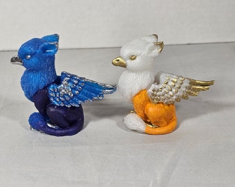 May include: Two small, decorative griffin figurines. One is blue with silver accents, the other is white with gold accents. Both have bird-like heads, feathered wings, and the bodies of a lion. They are sitting on a white surface.
