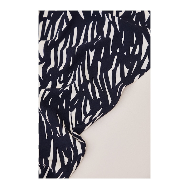 May include: Black and white abstract print fabric with a scribbled pattern. The fabric is folded and draped over a light beige surface.