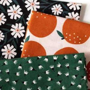 Könnte beinhalten: Drei gefaltete Stoffstücke mit verschiedenen Mustern. Eines ist schwarz mit weißen Blumen, ein anderes ist weiß mit Orangenfrüchten und das dritte ist grün mit weißen und schwarzen Blumenformen. Diese Stoffe sind wahrscheinlich zum Nähen oder Basteln.