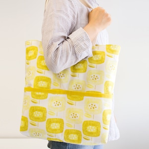 Könnte beinhalten: Eine weiße Tragetasche mit gelbem Blumenmuster. Die Tasche hat eine gelbe Stofftasche auf der Vorderseite und zwei lange Henkel.