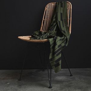 Könnte beinhalten: Ein grün-schwarzer gemusterter Schal, der über einen geflochtenen Stuhl mit schwarzen Metallbeinen gelegt ist. Der Schal trägt den Text "Find the Maker".