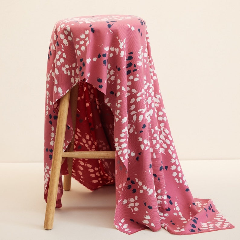 Pu&ograve; includere: Un tessuto rosa con una stampa astratta bianca e blu navy drappeggiato su uno sgabello di legno. Il tessuto ha una consistenza morbida e testurizzata.