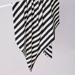 Może przedstawiać: Czarno-biała szalik lub szal w paski jest zawieszony na białym pręcie. Tkanina ma odważne, ukośne paski. Szalik jest wykonany z miękkiego materiału i wisi na gładkim, białym tle.