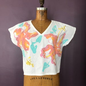 Könnte beinhalten: Weißes V-Ausschnitt-Oberteil mit kurzen Ärmeln, verziert mit großen, abstrakten floralen Designs in Rosa, Orange und Gelb, akzentuiert durch türkisfarbene und goldene Details. Das Oberteil wird auf einer Vintage-Schneiderpuppe präsentiert.