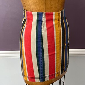 May include: A colorful, vertically striped mini skirt. The skirt features bold stripes of red, white, blue, yellow, green, and black. The garment is displayed on a wooden dress form, showcasing its form-fitting design.