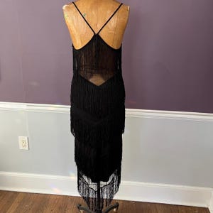 May include: A black sleeveless dress featuring multiple horizontal tiers of long fringe. The garment has thin spaghetti straps and a V-shaped back design, with the fringe extending from the bodice down to the hemline in a layered pattern.