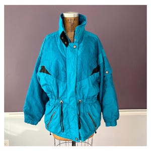 May include: A vintage teal ski jacket with black accents. The jacket features a high collar, black buttons, and a drawstring waist. It has two zippered pockets and black detailing on the shoulders. The jacket is displayed on a mannequin.