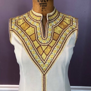 Puede incluir: Vestido sin mangas color crema con un escote bordado. El bordado presenta detalles dorados, burdeos y verdes en un patrón geométrico y floral. El vestido tiene un cuello alto y una línea vertical de bordado en la parte delantera.