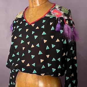 Pode incluir: Blusa preta de manga comprida com decote em V e um padrão de triângulos turquesa, laranja e brancos. Os ombros são adornados com penas e borlas roxas. O decote é adornado em vermelho.