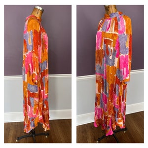 Puede incluir: Un vestido largo y fluido con cuello alto y mangas largas. El vestido presenta un vibrante estampado abstracto en tonos naranja, rosa, morado y blanco. El vestido se exhibe en un maniqu&iacute; vintage.