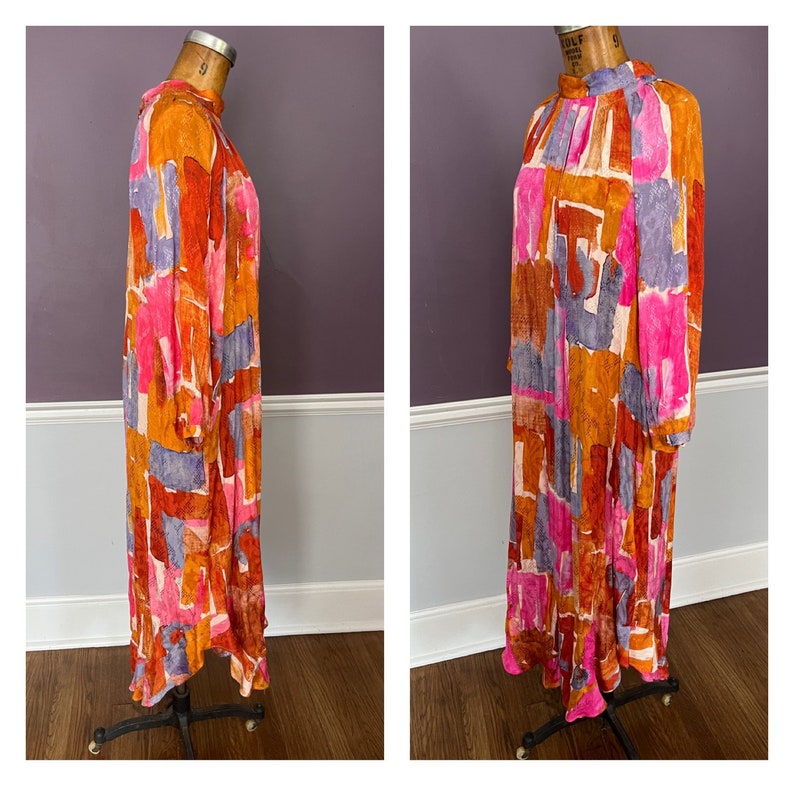 Puede incluir: Un vestido largo y fluido con cuello alto y mangas largas. El vestido presenta un vibrante estampado abstracto en tonos naranja, rosa, morado y blanco. El vestido se exhibe en un maniqu&iacute; vintage.