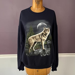 Pode incluir: Camisola azul marinho com um design gráfico detalhado de um lobo sob a lua cheia, rodeado por árvores. O design é realista. A camisola é exibida em um manequim vintage.