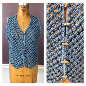May include: A light blue denim vest with a woven design. The vest has a V-neck and fastens with wooden toggle buttons. The sleeveless top is displayed on a vintage mannequin.