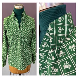 May include: A long-sleeved green shirt featuring a white grid pattern with small floral motifs in each square. The shirt has a pointed collar, a V-neckline, and a single button on the collar point. It is layered over a dark green turtleneck.