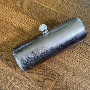 Puede incluir: Un clutch plateado con un acabado texturizado y un cierre con incrustaciones de cristal. El bolso cilíndrico está hecho de un material metálico y descansa sobre una superficie de madera. El clutch es un accesorio elegante.