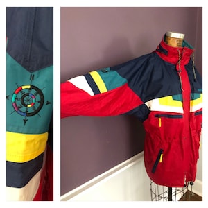 Op de afbeelding: Een kleurrijke vintage jas met een nautisch thema. De jas heeft een rode basis met marineblauwe, teal, gele en witte accenten. Een kompasroos is op de mouw geborduurd. De jas heeft een capuchon en meerdere zakken.