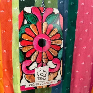 May include: A colorful tag with an embroidered floral design hangs against a striped fabric background. The tag features a large, vibrant flower in shades of red, orange, and pink, with green leaves. The tag reads "FARM RIO. DE. JANEIRO".