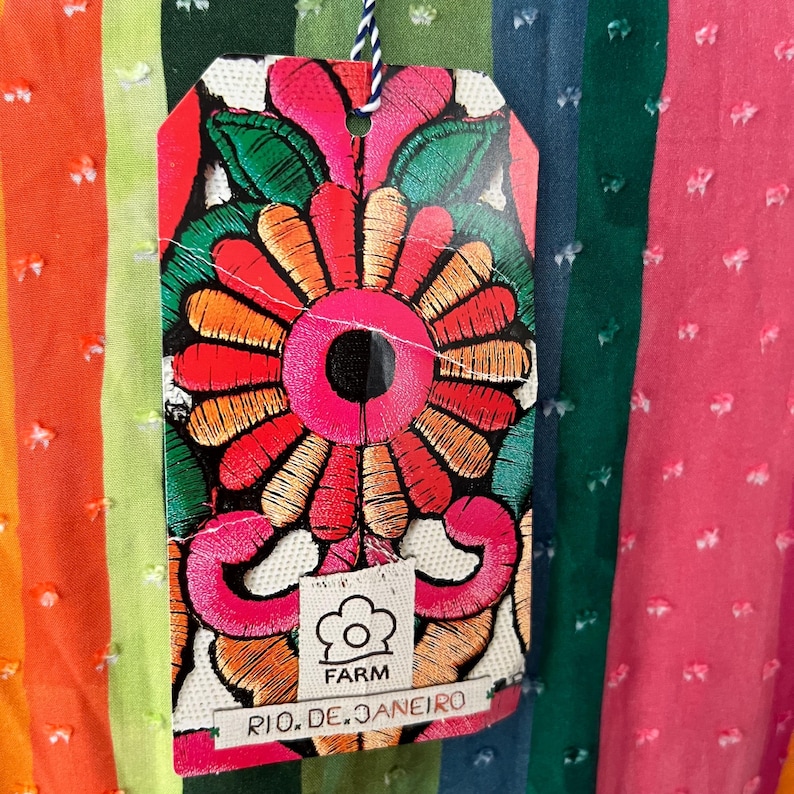 May include: A colorful tag with an embroidered floral design hangs against a striped fabric background. The tag features a large, vibrant flower in shades of red, orange, and pink, with green leaves. The tag reads "FARM RIO. DE. JANEIRO".