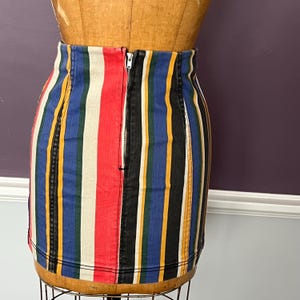 May include: A denim mini skirt with vertical stripes in red, white, blue, yellow, and black. The skirt has a front zipper closure and is displayed on a mannequin.
