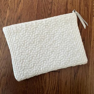 Könnte beinhalten: Eine weiße gewebte Clutch mit rechteckiger Form. Die Clutch hat eine strukturierte Oberfläche mit einem Korbwebmuster. Ein kleiner, hellfarbener Lederriemen ist an der oberen Ecke befestigt. Die Clutch liegt auf einer Holzoberfläche.