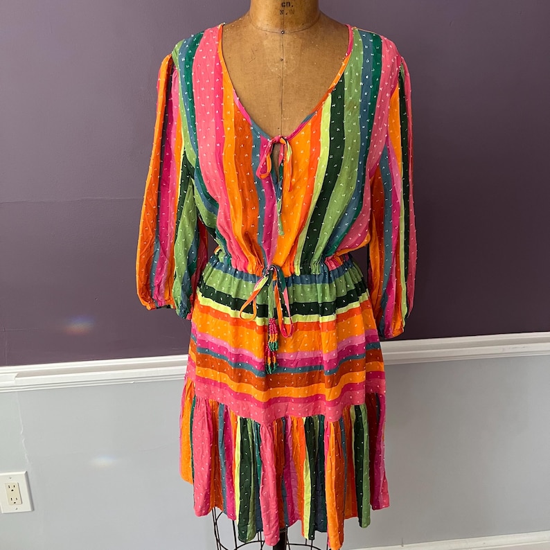 May include: A colorful, striped dress with long sleeves and a V-neck. The dress features vertical stripes in shades of pink, orange, green, and black. It has a drawstring waist and a ruffled hem. The dress is displayed on a vintage dress form.