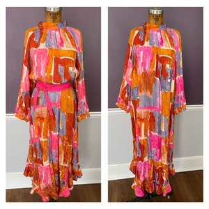 Puede incluir: Un vestido de manga larga con cuello alto y cintur&oacute;n rosa brillante. El vestido presenta un colorido estampado abstracto en tonos naranja, rosa, morado y marr&oacute;n. El vestido tiene una falda fluida y se exhibe en un maniqu&iacute;.