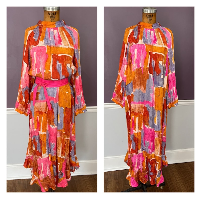 Puede incluir: Un vestido de manga larga con cuello alto y cintur&oacute;n rosa brillante. El vestido presenta un colorido estampado abstracto en tonos naranja, rosa, morado y marr&oacute;n. El vestido tiene una falda fluida y se exhibe en un maniqu&iacute;.