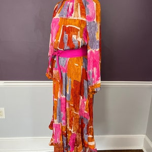 Puede incluir: Vestido de manga larga con cuello alto y un vibrante estampado abstracto en tonos naranja, rosa y morado. El vestido est&aacute; ce&ntilde;ido a la cintura con un cintur&oacute;n rosa brillante. El vestido se exhibe en un maniqu&iacute;.