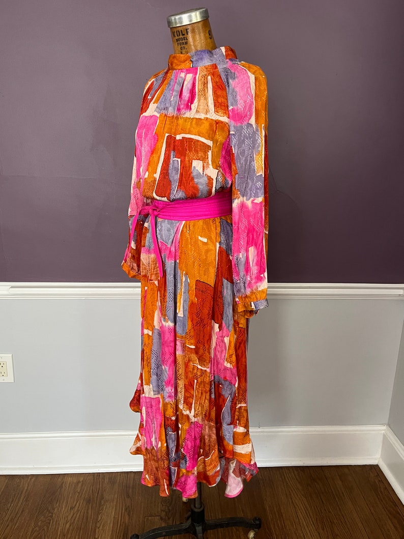 Puede incluir: Vestido de manga larga con cuello alto y un vibrante estampado abstracto en tonos naranja, rosa y morado. El vestido est&aacute; ce&ntilde;ido a la cintura con un cintur&oacute;n rosa brillante. El vestido se exhibe en un maniqu&iacute;.