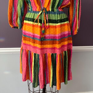 May include: A colorful, striped dress with long sleeves and a tiered skirt. The dress features a variety of bright colors, including orange, pink, green, and blue. It has a drawstring waist and a beaded tassel detail. The dress is displayed on a dress form.