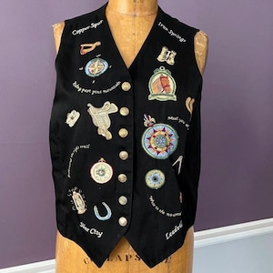 May include: A black vest featuring embroidered western-themed motifs including a saddle, compass, binoculars, lantern, and horseshoe. The garment includes gold-toned buttons and text such as Copper Spur, Iron Springs, and Silver City embroidered in white.