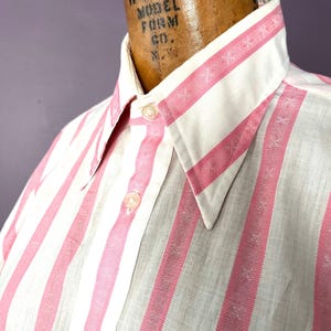 May include: Close-up of a vintage button-down shirt with vertical pink and white stripes. The collar and cuffs feature a detailed pattern. The shirt is buttoned up and displayed on a mannequin.