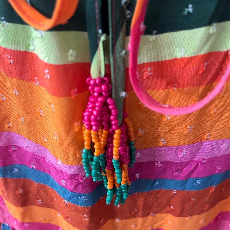 May include: Close-up of a colorful textile with horizontal stripes in shades of orange, pink, green, and blue. A beaded tassel in pink, orange, and green hangs from a green ribbon. The fabric has a textured, dotted pattern.