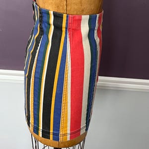 May include: A colorful striped skirt with vertical stripes in black, blue, yellow, red, and white. The skirt is made of a denim-like material and is displayed on a mannequin. The skirt has a classic, casual style.