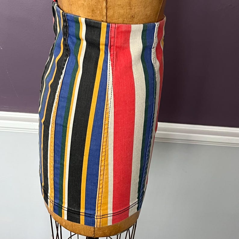 May include: A colorful striped skirt with vertical stripes in black, blue, yellow, red, and white. The skirt is made of a denim-like material and is displayed on a mannequin. The skirt has a classic, casual style.