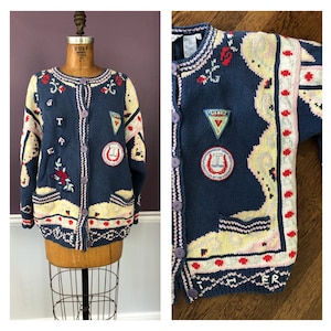 May include: Navy blue cardigan sweater with a button-up front. The sweater features embroidered floral and geometric patterns in red, white, and yellow. Patches with text and a horseshoe design are also present. The sweater is displayed on a mannequin.