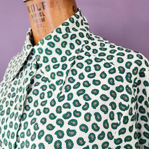 Pode incluir: Close-up de uma camisa branca abotoada com um padrão paisley verde e verde-azulado. A camisa está em um manequim de madeira. A gola e os detalhes dos botões são visíveis. A palavra "WOLF" é visível no manequim.