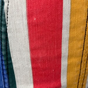 May include: Close-up of a section of striped fabric. The stripes are vertical and in the colors red, white, yellow, green, and blue. The fabric has a textured appearance, and the seams are visible on the left and right sides.