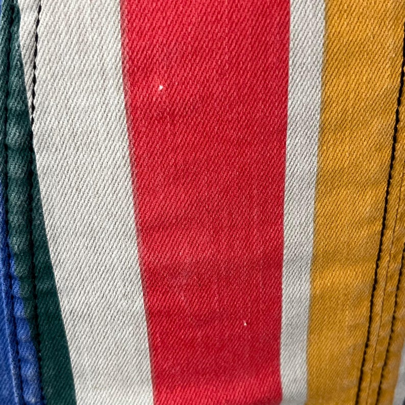 May include: Close-up of a section of striped fabric. The stripes are vertical and in the colors red, white, yellow, green, and blue. The fabric has a textured appearance, and the seams are visible on the left and right sides.