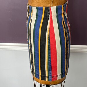 May include: A denim skirt with vertical stripes in red, white, blue, yellow, black, and green. The skirt is displayed on a mannequin. The skirt has a classic, straight cut and is made of a sturdy denim material.