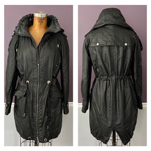 May include: Black hooded parka with a zip-up front, multiple pockets, and adjustable waist. The jacket features a high collar and silver-toned hardware. The coat is displayed on a vintage mannequin.
