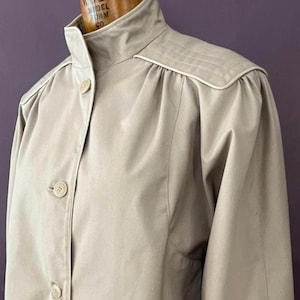 May include: A tan-colored vintage coat with a high collar and button-down front. The coat features shoulder pads and decorative stitching along the shoulders and collar. The coat is made of a smooth, possibly synthetic fabric.