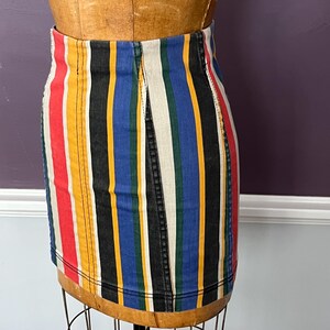 May include: A colorful, striped denim skirt displayed on a mannequin. The skirt features vertical stripes in red, yellow, black, blue, and white. The skirt is a mini-skirt style with a high waist and a classic denim design.